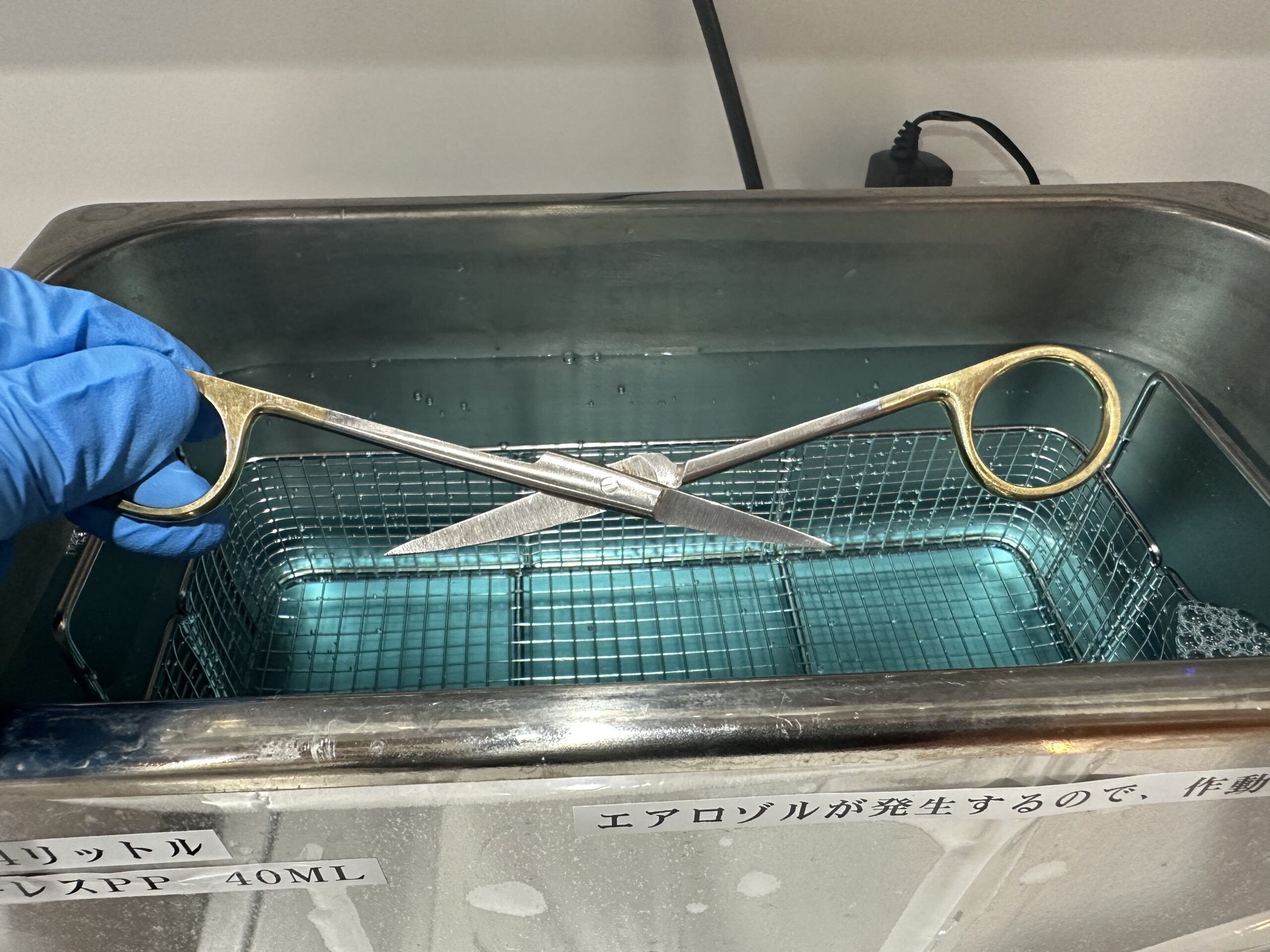 超音波洗浄器の網目のバスケットに、手袋をした手で器材を入れているところ。器材は洗浄液の中に沈める。