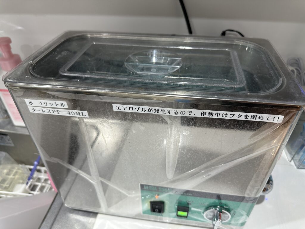 蓋を閉めた超音波洗浄器。本体に「水 4リットル」「エアロゾルが発生するので、作動中はフタを閉めて」と書いた紙が貼ってある。